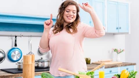 cheerful overweight woman listening music in headphones and dancing at table with fresh vegetables in kitchen at home